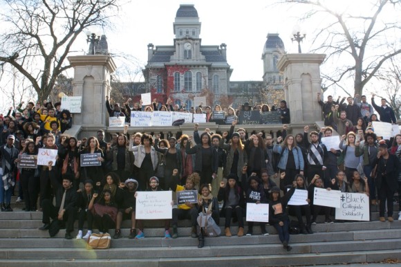 Photo of Syracuse University students standing on the steps outside of an academic building with their fists in the air or holding signs in protests.